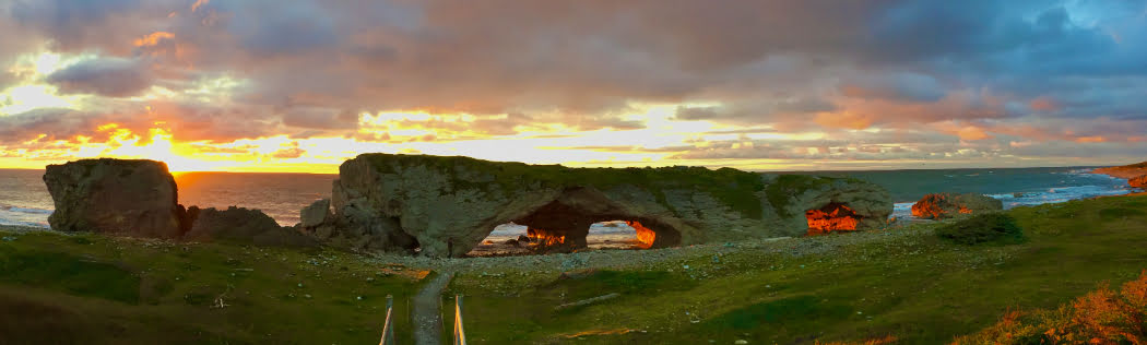 A grassy green space with large rocks and tunnels, with a body of water behind them, and a cloudy sunset.