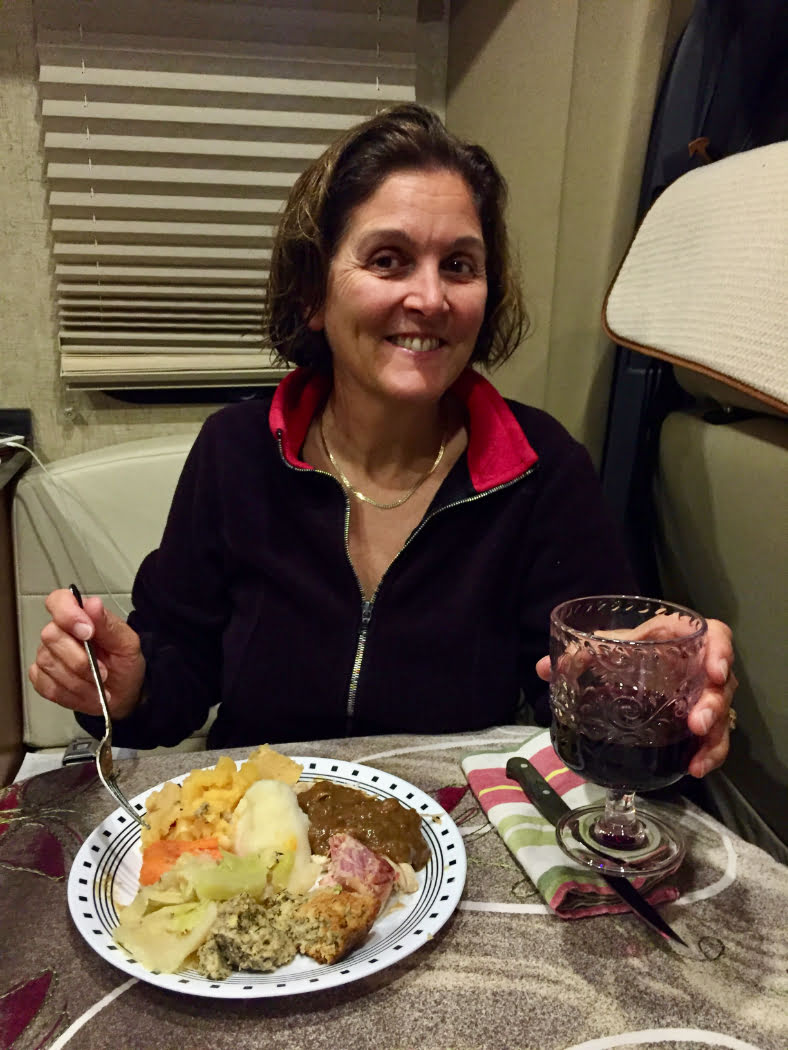 A woman in a black sweater smiles into the camera. She is holding a glass of red hand in her left hand and a fork in her right. In front of her is a plate full of food.