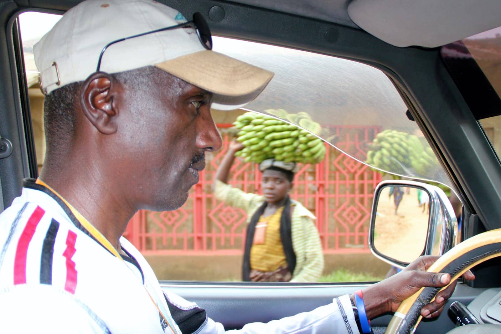 African man driving car in foreground with person carrying bananas in background