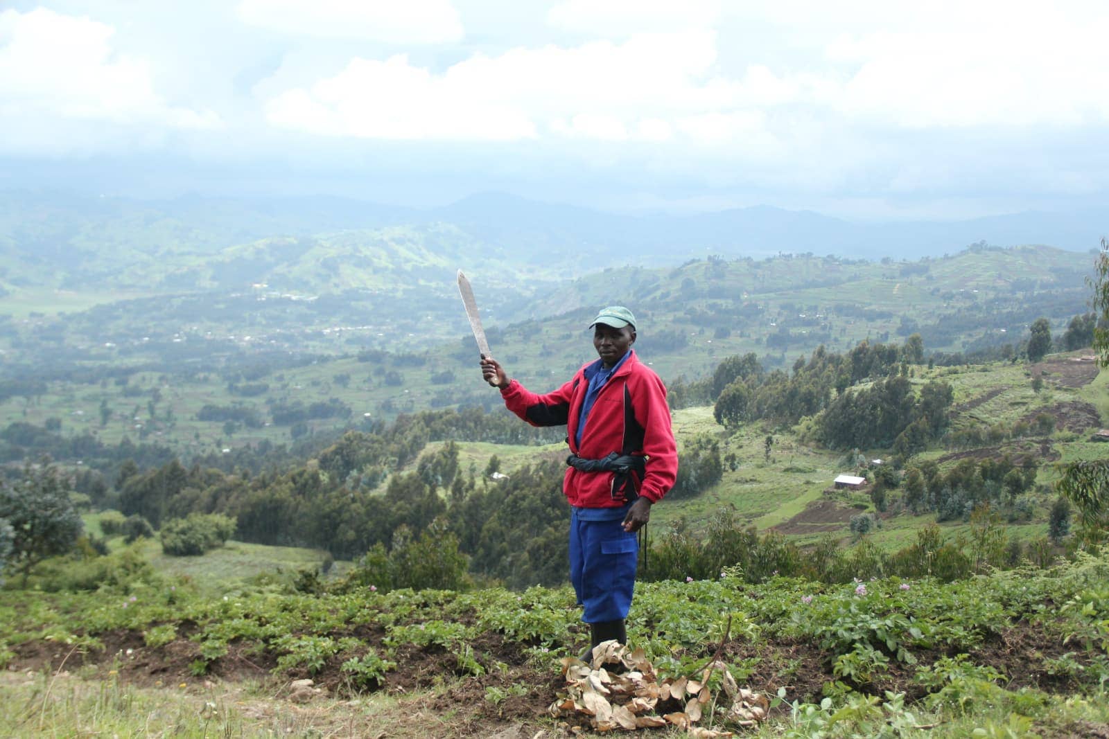 African man- n red jacket holding machete in foreground with green valley in background