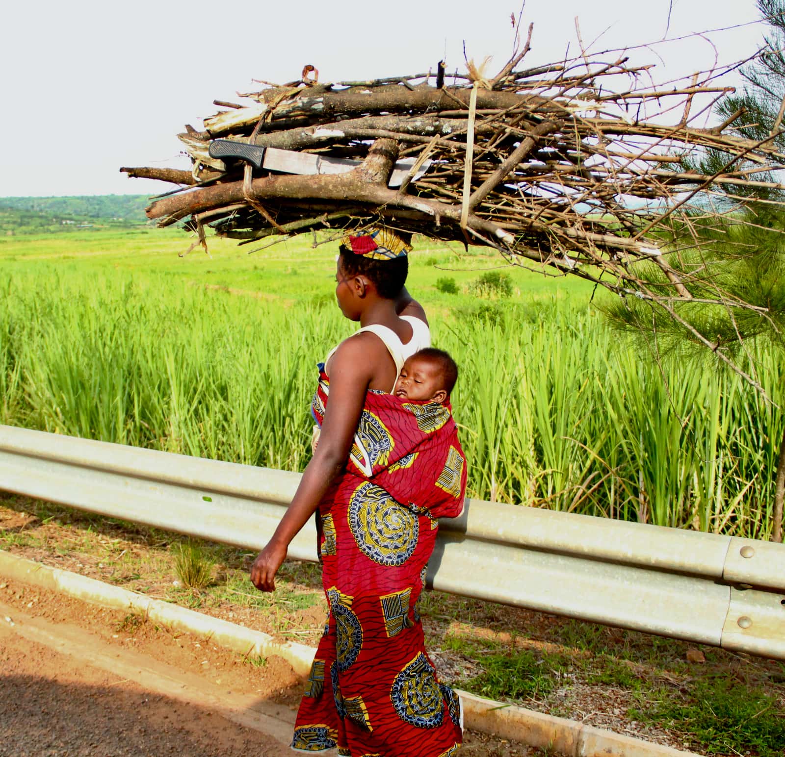 African woman with child on her back and sticks over her head walking on roadway