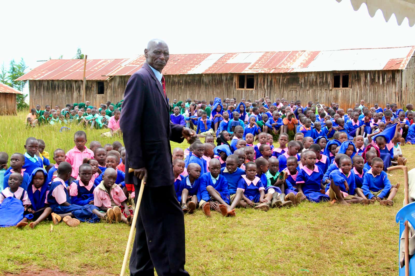 An elderly African man in a suit with dozens of children in blue school uniforms behind him