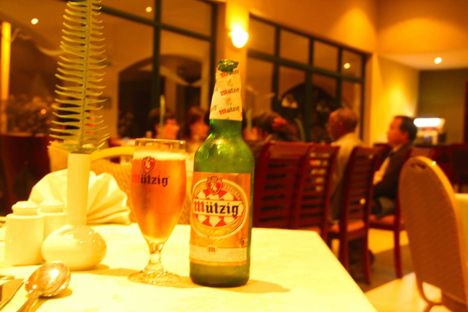 Beer bottle and glass in foreground with diners at restaurant in background