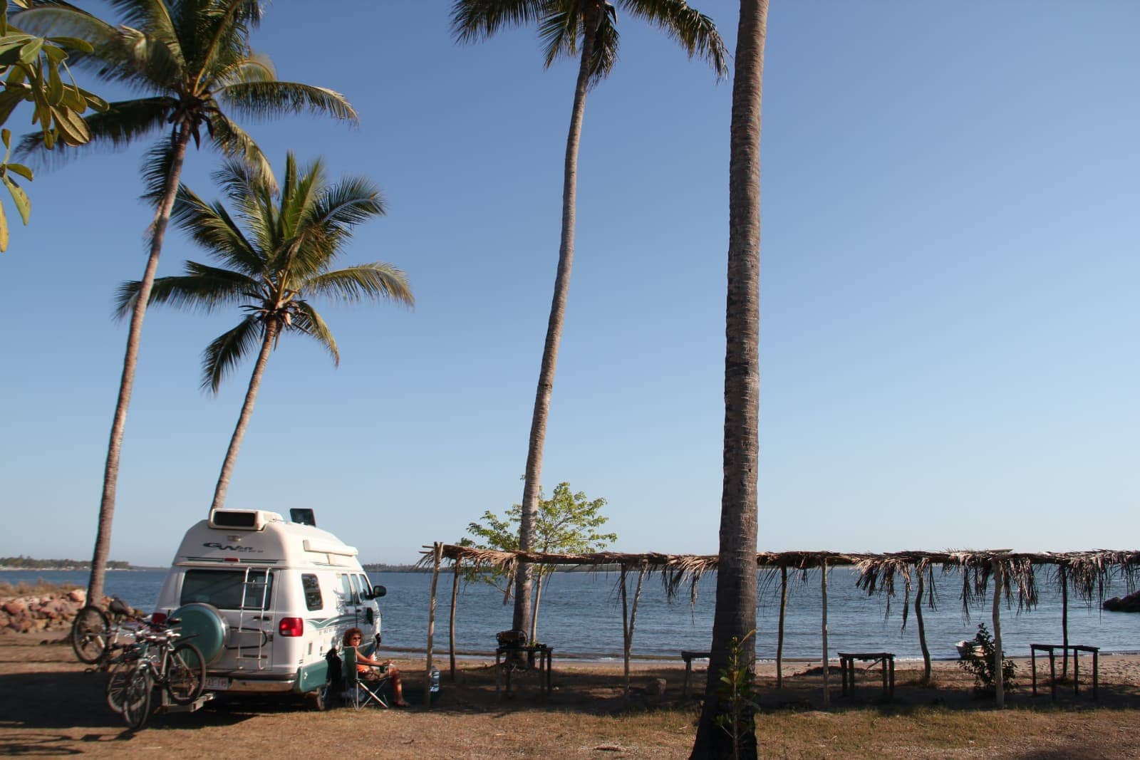 Camper van parked on beach with thatched roof shelter next to it