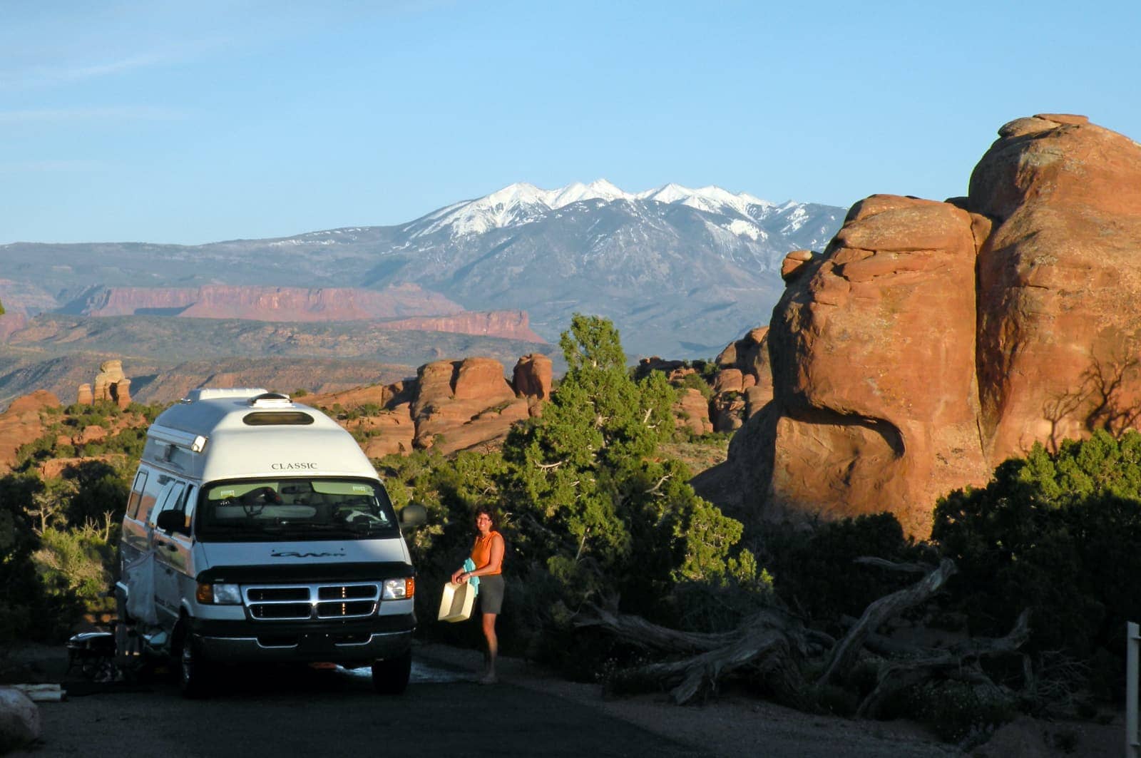 Woman and white camper van in foreground with mountains and blue sky in background