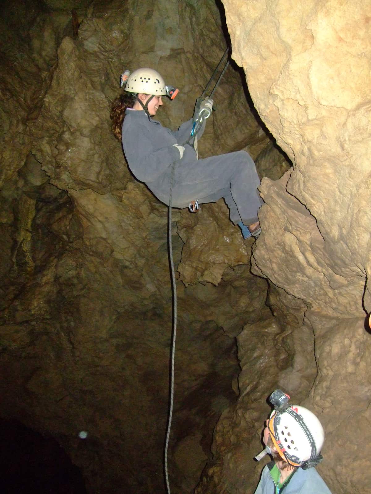 Woman in grey coveralls descending with rope in cave