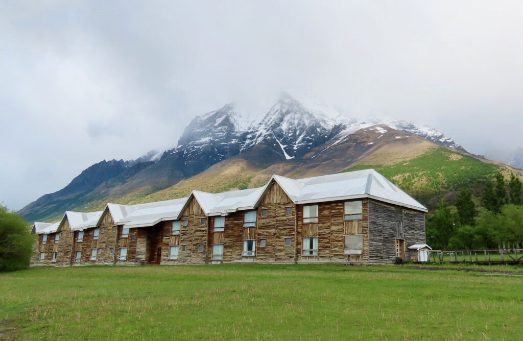 A long rustic wooden hotel with white metal rooflines sits on a green lawn, dwarfed by a snow capped mountain rising steeply behind it under cloudy skies.