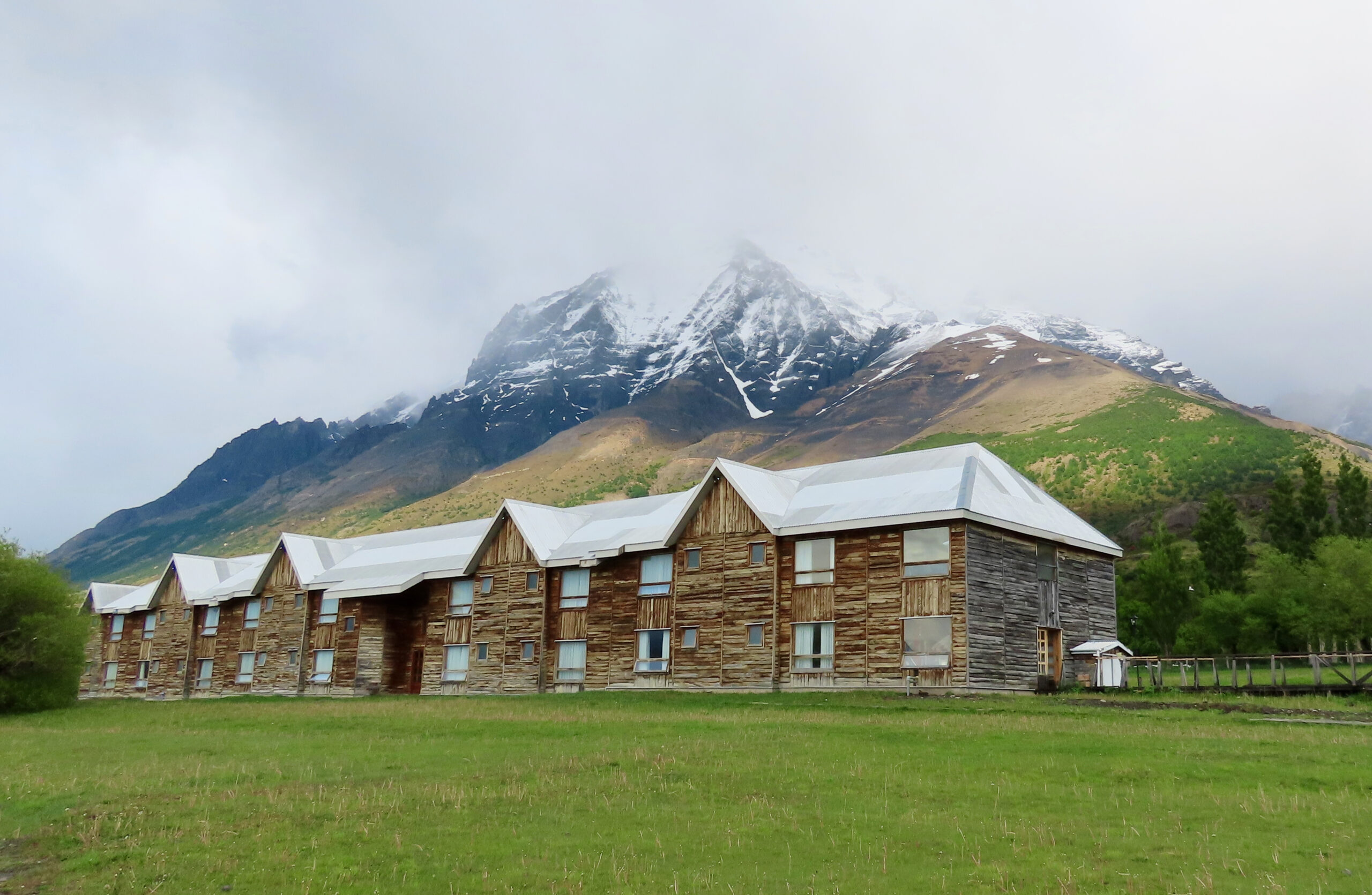 A long rustic wooden hotel with white metal rooflines sits on a green lawn, dwarfed by a snow capped mountain rising steeply behind it under cloudy skies.