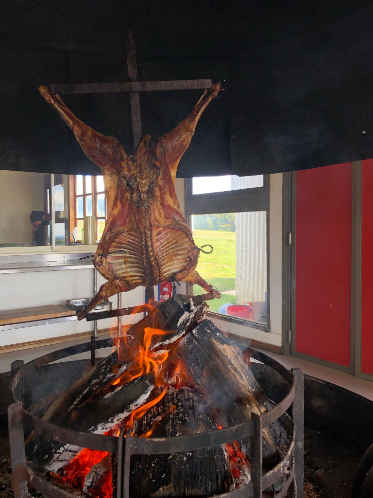 A whole lamb carcass mounted on a metal cross, roasting over an open wood fire in a large circular pit indoors.