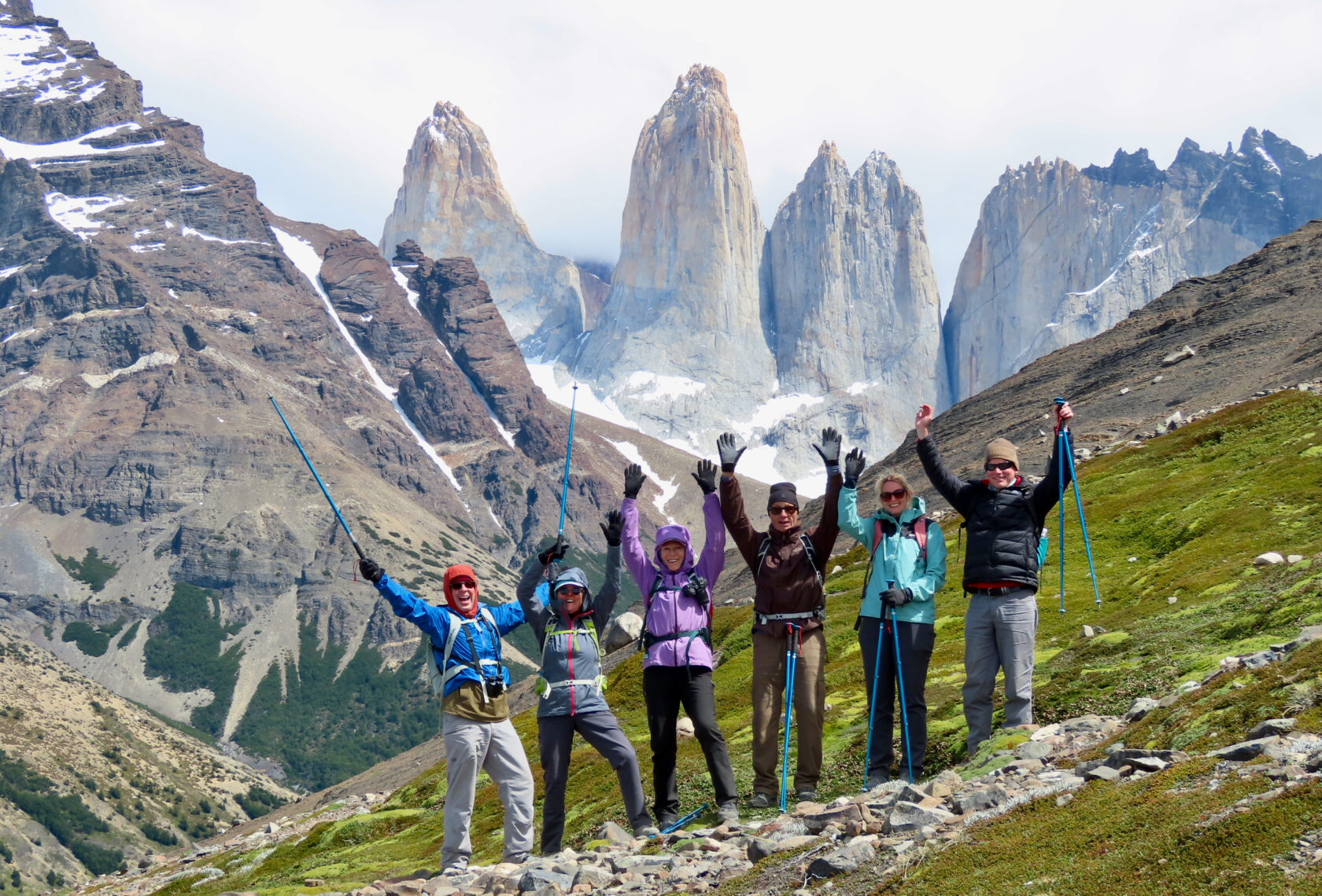 Six hikers in colourful gear with arms raised on a rocky hillside, with the dramatic granite towers of Torres del Paine rising behind them.