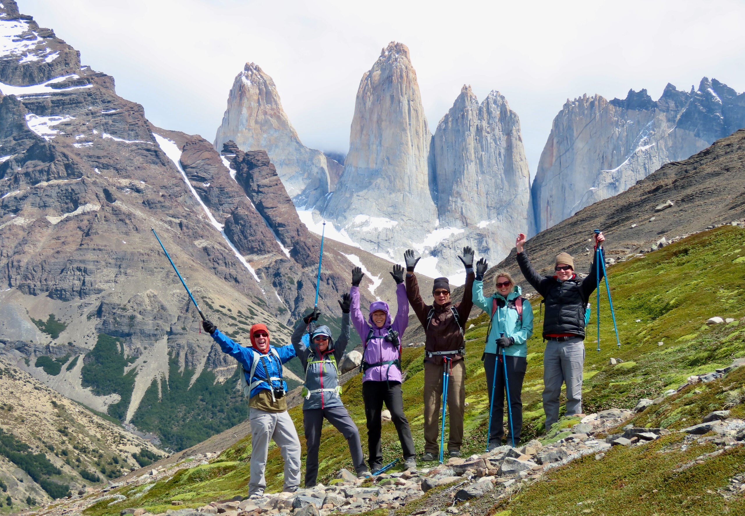 Six hikers in colourful gear with arms raised on a rocky hillside, with the dramatic granite towers of Torres del Paine rising behind them.