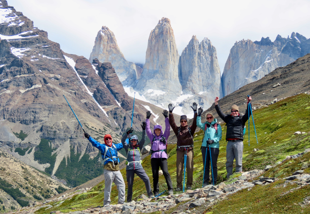 Six hikers in colourful gear with arms raised on a rocky hillside, with the dramatic granite towers of Torres del Paine rising behind them.