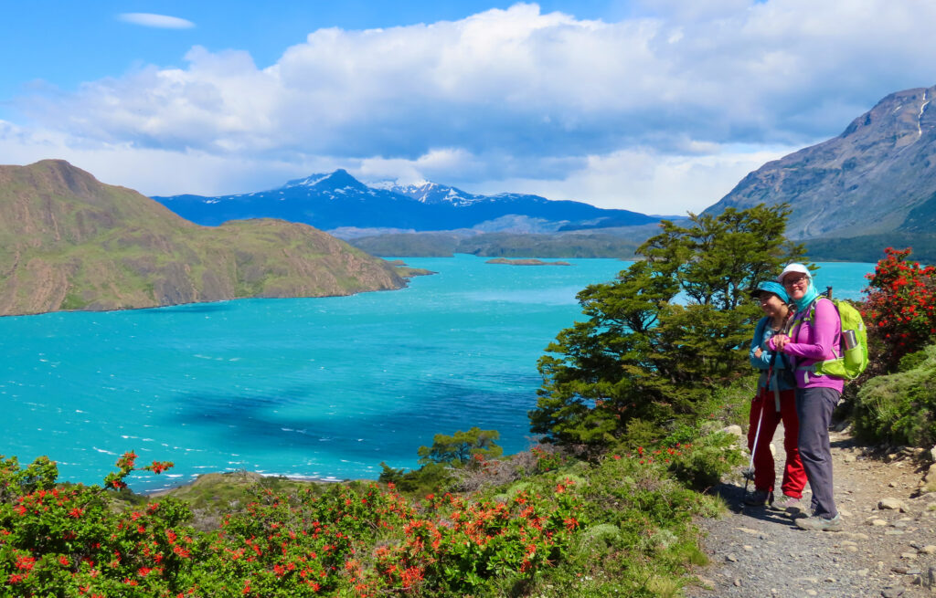 Two hikers pause on a trail above a vivid turquoise lake, with red-flowering shrubs in the foreground and snow capped mountains across the water.